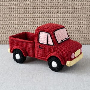 Könnte beinhalten: Ein roter gehäkelter Pickup-Truck mit hellrosa Windschutzscheibe und Seitenfenstern, einem cremefarbenen vorderen Stoßfänger und Kühlergrill sowie schwarzen Rädern mit weißen Zentren. Der Truck hat eine hohle Ladefläche und sichtbare Maschen.