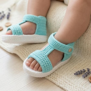 May include: Crocheted baby sandals in a light blue color with white soles. The sandals have a T-strap design and a strap that fastens with a button. The sandals are shown on a soft, cream-colored blanket.