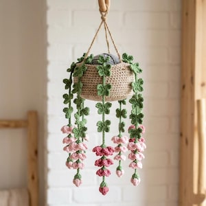 May include: A crocheted hanging plant in a beige basket with green vines and pink flowers. The plant features multiple long, trailing stems adorned with small green leaves and clusters of pink blossoms at the ends, suspended by a thin twine cord.
