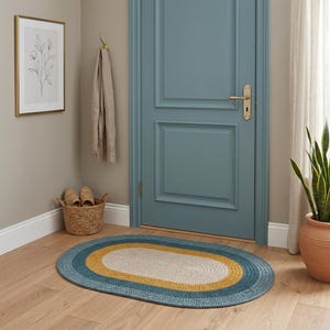 May include: An oval braided rug with a blue, yellow, and cream color scheme. The rug is placed on a hardwood floor in front of a blue door. A woven basket with slippers and a framed botanical print are also visible.