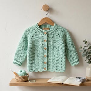 May include: A light green, crocheted baby cardigan with a scalloped hem and wooden buttons. The long-sleeved sweater is displayed on a wooden hanger. A small basket with yarn and a crochet hook sits on a shelf below.