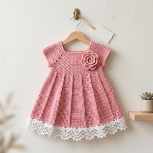 May include: A pink crochet dress with a white lace trim, short sleeves, and a square neckline. A crocheted flower is attached to the bodice. The dress is hanging on a wooden hanger.