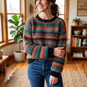 May include: A long-sleeved crochet sweater featuring a multi-colored granny square pattern in shades of teal, orange, purple, and grey. The garment has a solid black ribbed crew neckline and black ribbed cuffs, paired with blue denim jeans.