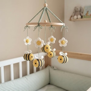 May include: A hanging mobile featuring crocheted bumblebees with yellow and gray stripes and white wings, interspersed with white crocheted flowers with yellow centers. The elements are suspended from a circular wooden frame by green yarn.