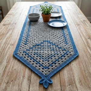 May include: A rectangular crochet table runner featuring a grey and blue granny square pattern with a solid blue border and a decorative leaf-shaped tassel at the pointed end. The runner is displayed on a wooden surface with a small potted plant.