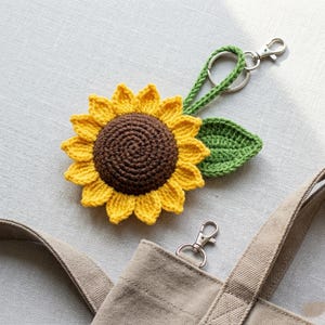 May include: A crochet sunflower keychain featuring a dark brown center, bright yellow petals, and a green leaf. The sunflower is attached to a green crocheted loop and a silver metal lobster clasp, which is clipped to a beige canvas bag.