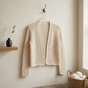 May include: A cream-coloured, open-front cardigan hangs on a clear hanger. The cardigan is made of a loosely woven knit fabric. The sleeves are long, and the garment has a simple, classic design. The background is a neutral wall with a small shelf and decorative items.