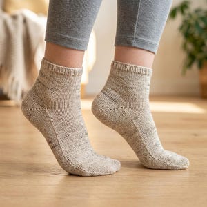 May include: A pair of ankle-length knit socks in a light beige color with subtle dark flecks throughout the fabric. The socks feature a ribbed cuff at the top and a visible knit texture, covering the feet up to the lower ankle area.