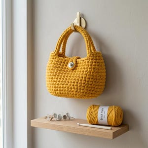 May include: A mustard yellow crochet handbag with a silver clasp hangs from a brass hook. Below, a wooden shelf holds a ball of "Brick Yarn" and a crochet hook. The bag has two handles and a rounded shape.