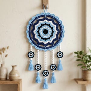 May include: A circular wall hanging featuring a crocheted mandala in shades of navy blue, light blue, white, and black. The piece includes five hanging strands decorated with white beads, small crocheted flower motifs, and light blue tassels.