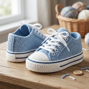 May include: A pair of baby blue crochet sneakers with white laces and a white toe cap. The sneakers have a white sole with a blue stripe. The shoes are sitting on a wooden surface.