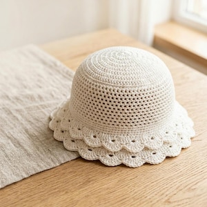 May include: A cream-coloured crochet hat with a scalloped brim sits on a wooden surface. The hat has a rounded crown and a wide, decorative brim. The open weave design allows for breathability, suitable for warm weather.