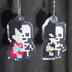 May include: Two clear acrylic keychains hanging from silver ball chains. The left keychain features a pixel art design of a figure with a red cape and a skeleton half. The right keychain shows a pixel art figure in a dark suit with a skeleton half.