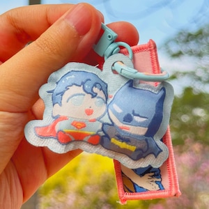 May include: A keychain featuring cartoon depictions of Superman and Batman. The characters are in a cute, chibi style, with Superman in his classic red and blue suit and Batman in a blue and black costume. The keychain has a light blue clasp and a pink strap.