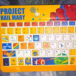 May include: A yellow wireless keyboard featuring the text Project Hail Mary at the top left. The keyboard has a colorful layout with various space-themed illustrations on the keys, including planets, a rocket, and a fox, alongside standard QWERTY lettering.