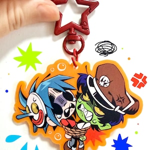 Puede incluir: Un llavero de acr&iacute;lico de doble cara con un cierre en forma de estrella roja. El dije muestra una ilustraci&oacute;n de dibujos animados de un personaje de pelo azul con una m&aacute;scara blanca y un personaje de piel verde con un sombrero marr&oacute;n y s&iacute;mbolos.