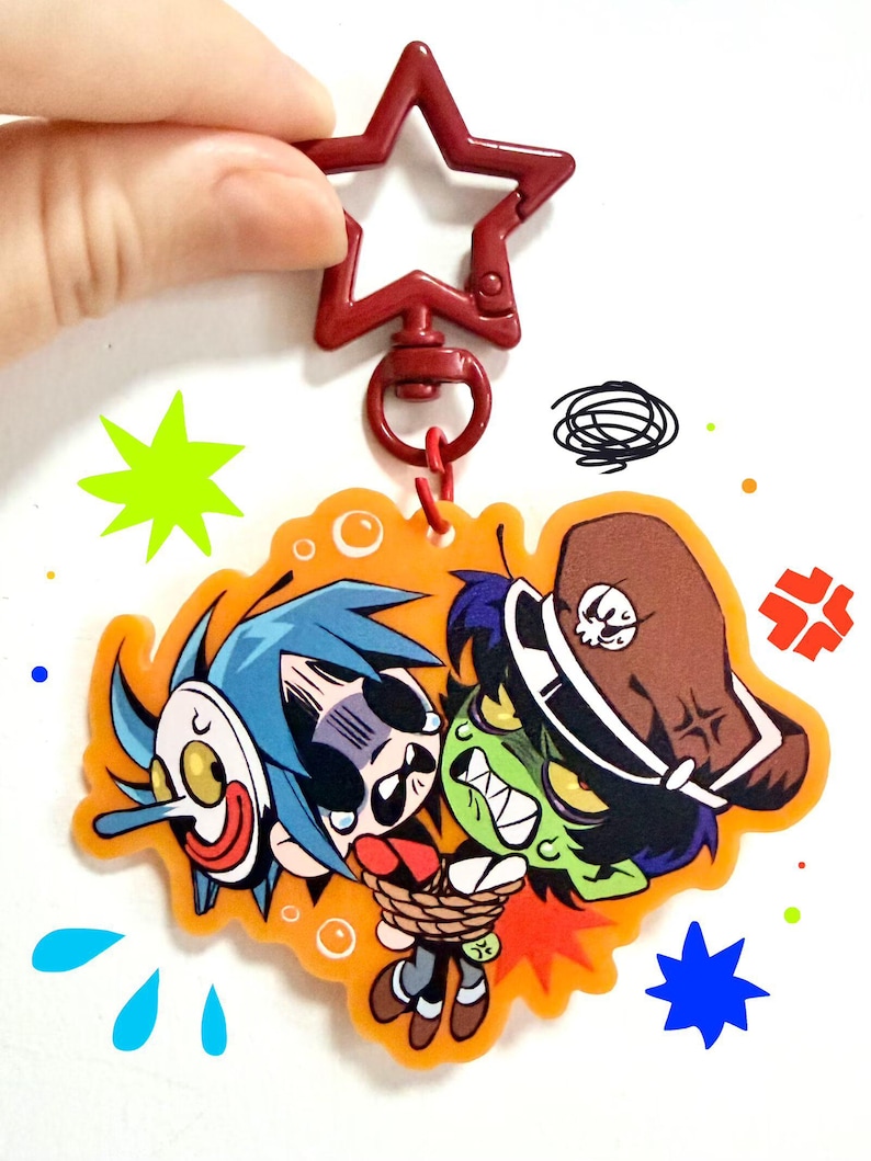 Puede incluir: Un llavero de acr&iacute;lico de doble cara con un cierre en forma de estrella roja. El dije muestra una ilustraci&oacute;n de dibujos animados de un personaje de pelo azul con una m&aacute;scara blanca y un personaje de piel verde con un sombrero marr&oacute;n y s&iacute;mbolos.