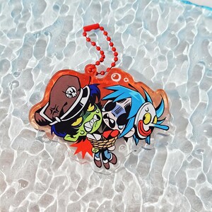 Puede incluir: Un llavero de acr&iacute;lico transparente con una ilustraci&oacute;n de dibujos animados de un personaje de piel verde con sombrero de pirata marr&oacute;n que sostiene a un personaje de pelo azul atado con una cuerda y una m&aacute;scara blanca de nariz larga.