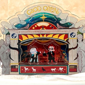 May include: A multi-layered acrylic display featuring characters from "Good Omens." The design includes a stage with two figures, angels, and the text "GOOD OMENS" across the top. The color palette includes blue, red, yellow, and white.