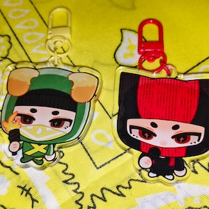 Puede incluir: Dos llaveros acrílicos con diseños de personajes de dibujos animados. Uno presenta un atuendo verde con una antorcha, el otro un atuendo rojo y negro. Ambos tienen cierres dorados o rojos y están sobre un fondo amarillo estampado.