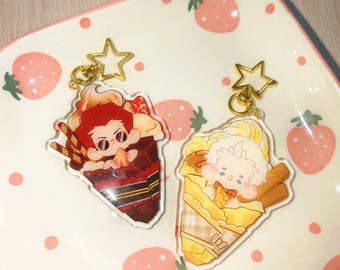 May include: Two acrylic keychains featuring cartoon characters in dessert-themed designs. One keychain has a red-haired character in a chocolate dessert, and the other has a blonde character in a yellow dessert. Both keychains have gold star-shaped clasps.
