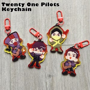 Puede incluir: Cuatro llaveros de Twenty One Pilots con cierres rojos. Cada dije presenta un personaje de dibujos animados con varios atuendos, algunos sosteniendo instrumentos. El texto "Twenty One Pilots Keychain" está en la parte superior.
