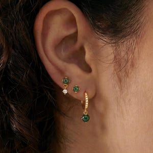 May include: A close-up of an ear adorned with three different gold earrings. One is a small stud with a white gem, another is a small stud with a green gem, and the third is a hoop earring with a green gem and small white gems.