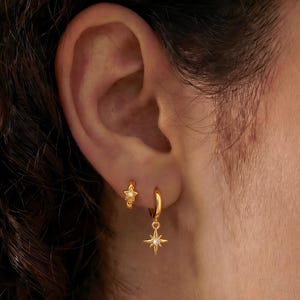 May include: Two gold hoop earrings worn in the earlobe. The smaller hoop features a tiny star-shaped charm with a central stone. The larger hoop hangs below it, featuring a dangling eight-pointed star charm with a small, clear, sparkling stone.