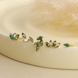 Op de afbeelding: Vijf goudkleurige oorknopjes genummerd van één tot vijf. Elke oorbel bevat marquise-geslepen mosagaatstenen in groen en wit, waarbij één oorbel een heldere steen bevat. De ontwerpen variëren in het aantal stenen en de rangschikking.