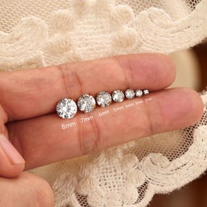 May include: A series of seven round, clear, faceted stud earrings arranged by size from 8mm to 2mm. Each earring features a brilliant-cut stone set in a metal base, with the corresponding metric size clearly labeled below each individual piece.