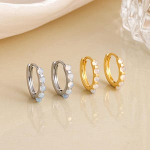 May include: Four hoop earrings, two silver and two gold, each adorned with small, light-colored gemstones. The earrings are displayed on a neutral background, showcasing their design and color variations.