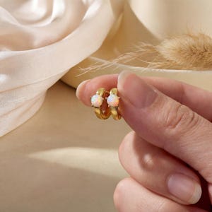 May include: A pair of gold hoop earrings, each featuring a round, iridescent opal stone. The earrings are held up against a soft, neutral-toned background, highlighting their delicate design and warm gold color.