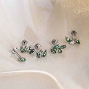 May include: An assortment of silver-toned earrings with a leaf design. Each earring features multiple green, teardrop-shaped stones. The earrings are displayed on a white surface, showcasing their delicate details and design.