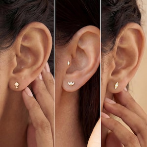 May include: Close-up of three gold stud earrings. The earrings feature different designs, including a small opal, a three-petal flower, and a small oval shape with small stones. The earrings are displayed on an ear.