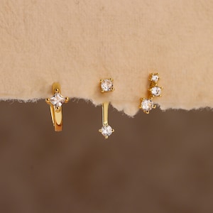 May include: A selection of gold-coloured earrings featuring clear cubic zirconia stones. The earrings include a small hoop with a square stone, a stud, a dangling earring, and a curved design with multiple stones. The earrings are displayed on a neutral background.