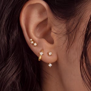 Pode incluir: Um conjunto de brincos dourados composto por um pequeno pino com três pedras, uma argola com uma pedra quadrada única e um brinco de duas partes com uma pedra quadrada na frente e uma pedra pendente na parte de trás.