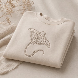 May include: A beige crewneck sweatshirt featuring a detailed brown embroidered design of a stingray. The stingray illustration incorporates celestial motifs including a crescent moon, a sun, and various small stars across its wings and body.