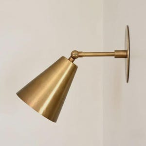 May include: A brass-toned wall-mounted sconce with a conical shade and adjustable arm. The sconce features a circular backplate and a pivoting joint for directional lighting. The fixture is mounted on a white wall.