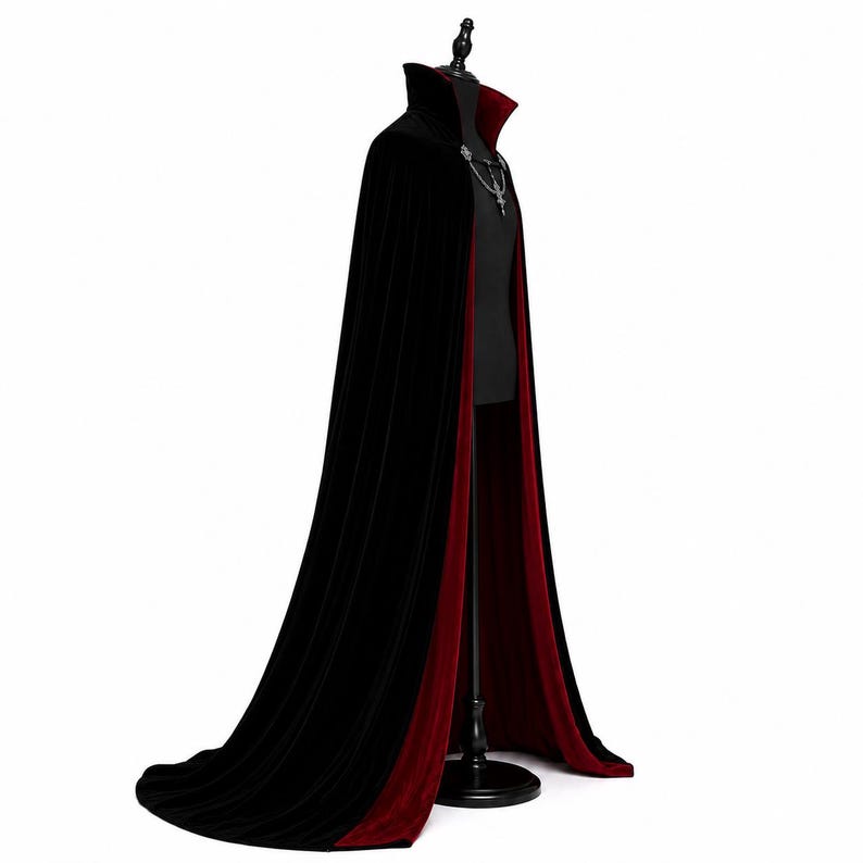 May include: A floor-length velvet cape featuring a deep black exterior and a contrasting dark red interior lining. The garment includes a high standing collar and a decorative silver-toned metal clasp at the neckline with a dangling chain detail.