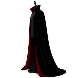 May include: A floor-length velvet cape featuring a deep black exterior and a contrasting dark red interior lining. The garment includes a high standing collar and a silver-toned metal clasp closure at the neckline, displayed on a mannequin.