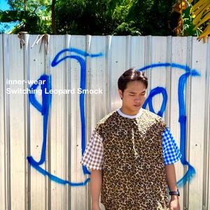 Puede incluir: Una blusa de manga corta con un panel central de estampado de leopardo, una manga de cuadros vichy marrón y blanco a la izquierda y una manga de cuadros vichy azul y blanco a la derecha. El texto "inner-wear Switching Leopard Smock" aparece a la izquierda.