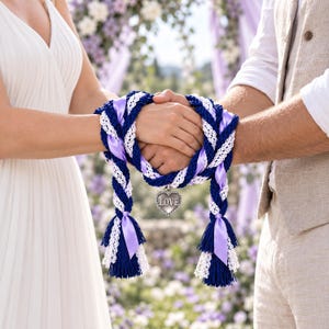 Könnte beinhalten: Ein Handfasting-Band aus geflochtenem marineblauem Seil, weißer Spitzenborte und lavendelfarbenem Satinband. Das Band ist um zwei ineinander verschränkte Hände gewickelt und mit einem silberfarbenen Herzanhänger mit der Aufschrift LOVE versehen.