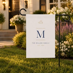 May include: A cream-coloured garden flag on a black metal stand. The flag displays a navy blue letter "M" and the words "THE MILLER FAMILY EST 2024" in a serif font. A small house icon is at the top.