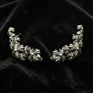 May include: A pair of silver ear climbers featuring a curved design composed of multiple interconnected cross motifs. Each earring displays a series of polished metallic floral-shaped crosses arranged in a graduated arc, creating a textured appearance.