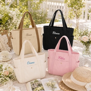 May include: Four canvas tote bags in beige, black, off-white, and light pink. Each bag features two top handles, side pockets, a front pocket with a gold snap button, and embroidered names: Briar, Elowen, Oriana, and Chloe in matching thread colors.