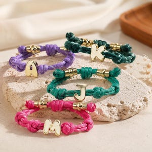 Peut inclure: Quatre bracelets en cordon réglable de couleurs violet, vert foncé, bleu sarcelle et rose vif. Chaque bracelet présente une breloque centrale en métal doré en forme de lettre, soit A, K, J ou M, ornée de petites perles métalliques dorées.
