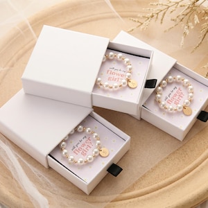 May include: Three white sliding gift boxes each containing a beaded bracelet made of white pearls and small gold-toned beads. Each bracelet features a round gold-toned charm with a letter and rests on a card printed with the text Will you be my flower girl?