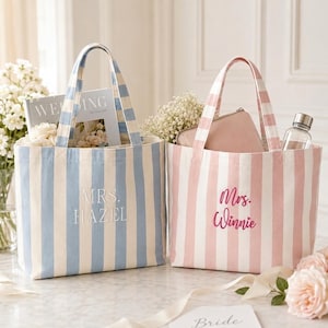 Pode incluir: Duas bolsas de lona com listras verticais. A bolsa da esquerda é azul clara e branca com o texto bordado MRS. HAZEL. A bolsa da direita é rosa clara e branca com o texto bordado Mrs. Winnie. Ambas as bolsas possuem duas alças superiores.