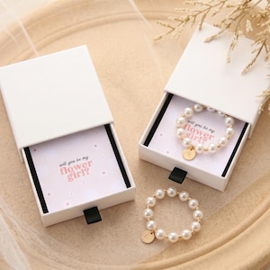 May include: Two white sliding gift boxes containing pearl beaded rings with small gold-toned circular charms. One ring features the letter R and the other the letter G. Each box includes a card with the text will you be my flower girl?