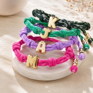 Op de afbeelding: Vier koordarmbanden in donkergroen, felgroen, paars en felroze. Elke armband is voorzien van een enkele letterbedel van goudkleurig metaal, namelijk K, J, A en M, met goudkleurige metalen eindkapjes aan de uiteinden van het koord.